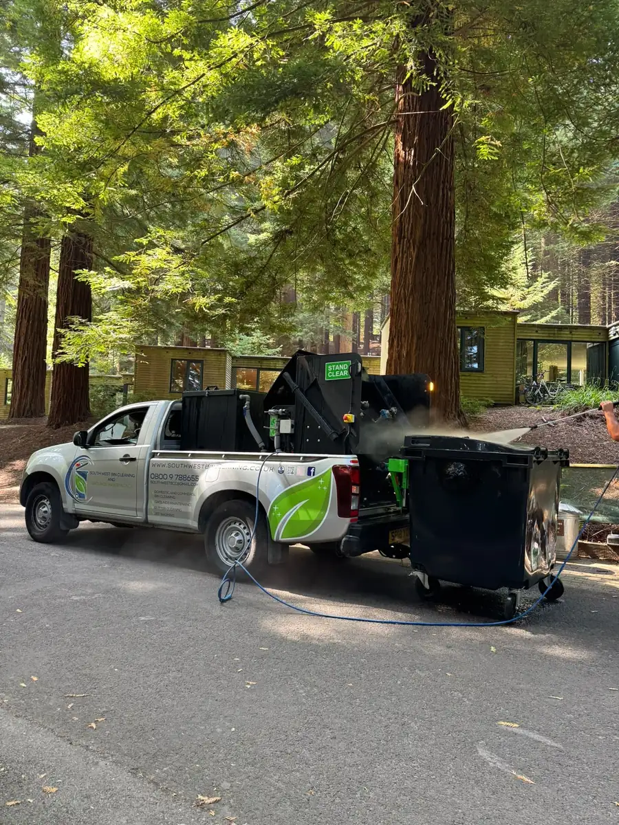 Commercial wheelie bin being washed on a pickup-mounted rig in a woodland setting